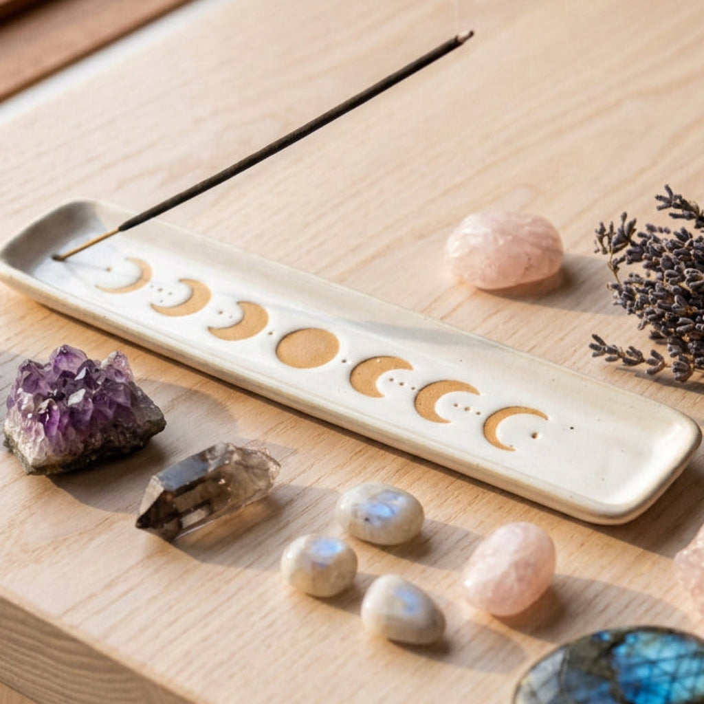 A white rectangular ceramic incense holder with a moon phase pattern, featuring a black incense stick resting diagonally across the top, set against a minimalist beige background.