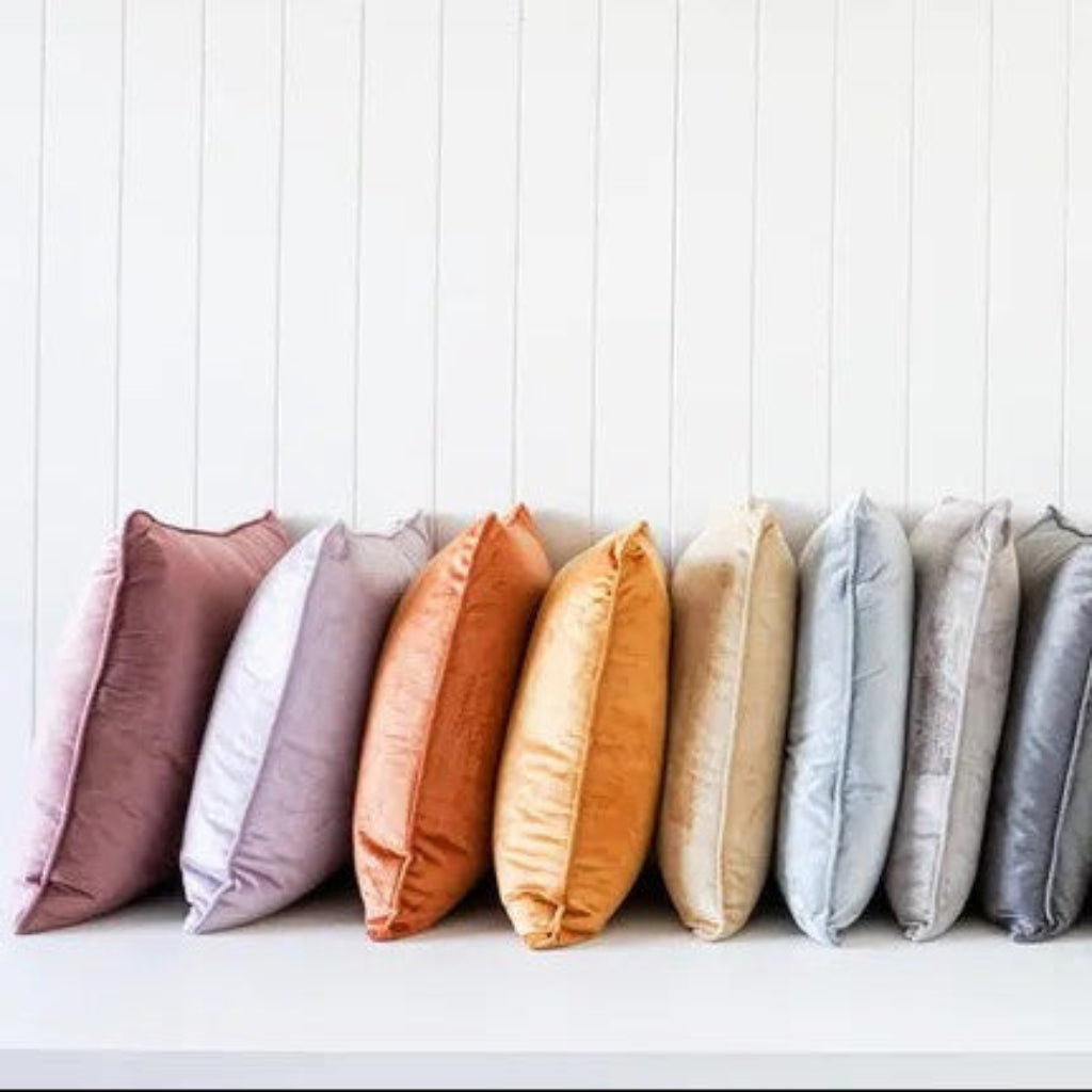 A row of twelve square throw pillows in a muted rainbow color palette, arranged neatly on a white shelf against a white vertical shiplap wall.