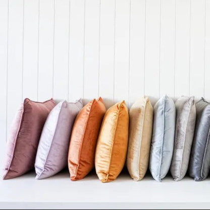 A row of twelve square throw pillows in a muted rainbow color palette, arranged neatly on a white shelf against a white vertical shiplap wall.