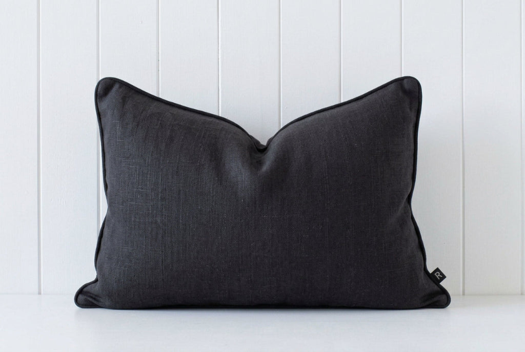 A rectangular black lumbar pillow with dark gray piping, resting against a white vertical wood paneled wall.