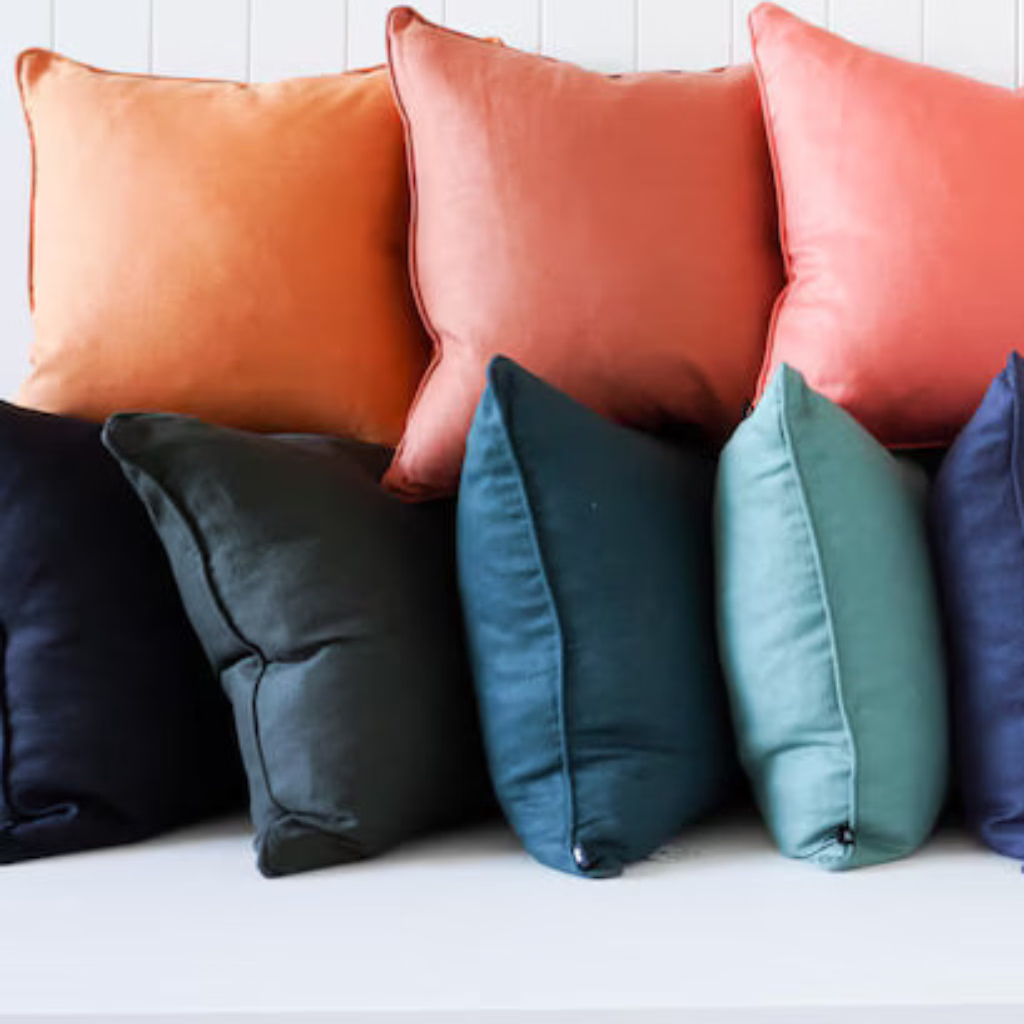 A row of square throw pillows in multiple colors, including orange, coral, pink, blush, teal, navy, gray, off-white, and cream, arranged on a white bench against a white paneled wall.