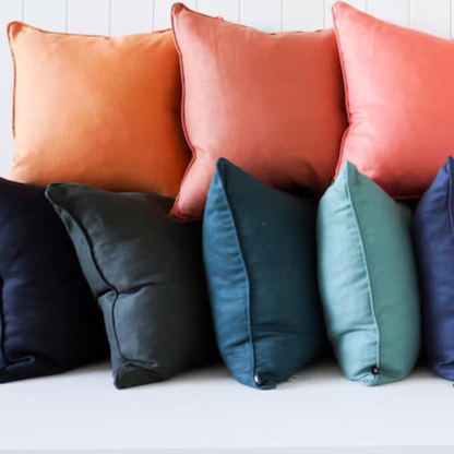 A row of square throw pillows in multiple colors, including orange, coral, pink, blush, teal, navy, gray, off-white, and cream, arranged on a white bench against a white paneled wall.