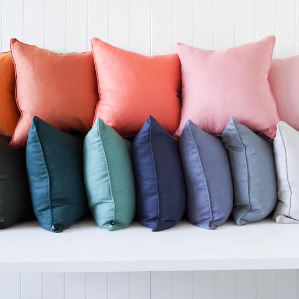 A row of square throw pillows in multiple colors, including orange, coral, pink, blush, teal, navy, gray, off-white, and cream, arranged on a white bench against a white paneled wall.