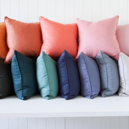 A row of square throw pillows in multiple colors, including orange, coral, pink, blush, teal, navy, gray, off-white, and cream, arranged on a white bench against a white paneled wall.