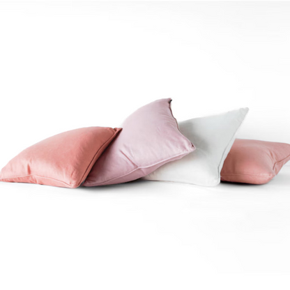 Four decorative throw pillows in dusty pink, light mauve, white, and medium pink, arranged in a pile on a seamless white studio background.