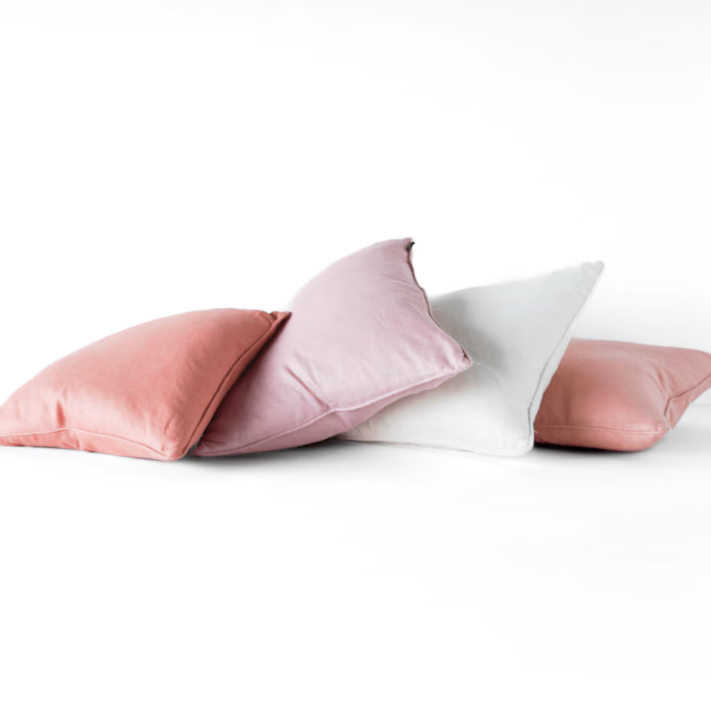 Four decorative throw pillows in dusty pink, light mauve, white, and medium pink, arranged in a pile on a seamless white studio background.