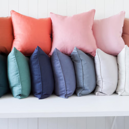 A row of square throw pillows in multiple colors, including orange, coral, pink, blush, teal, navy, gray, off-white, and cream, arranged on a white bench against a white paneled wall.