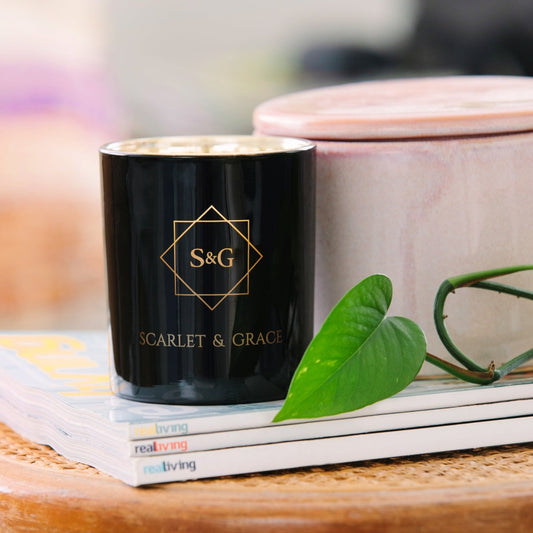 A black candle with a gold logo on the side, labeled 'Scarlet & Grace', placed on a table with a green leaf and a pink container in the background.